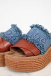 A pair of platform slide sandals featuring brown leather straps, a thick braided jute platform sole, and a frayed blue denim upper with the brand name 'Chloé' embroidered in denim.
