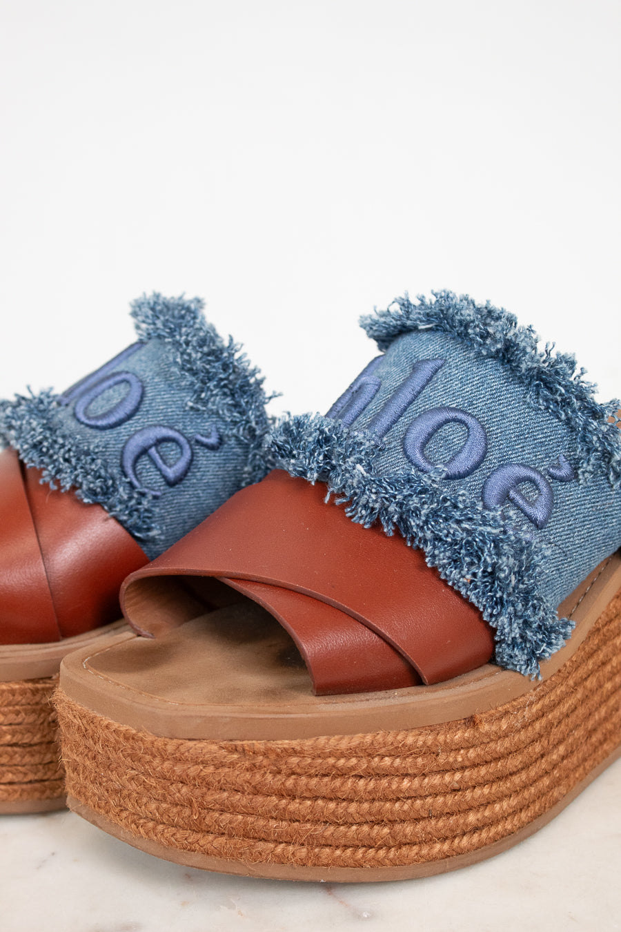 A pair of platform slide sandals featuring brown leather straps, a thick braided jute platform sole, and a frayed blue denim upper with the brand name 'Chloé' embroidered in denim.