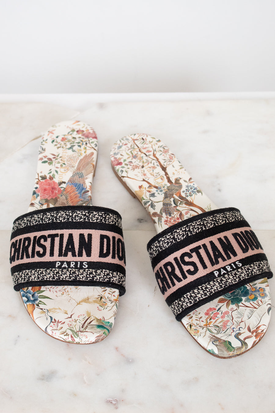 Pair of floral sandals with 'Christian Dior' woven into black/white/pink strap on a marble surface and white background