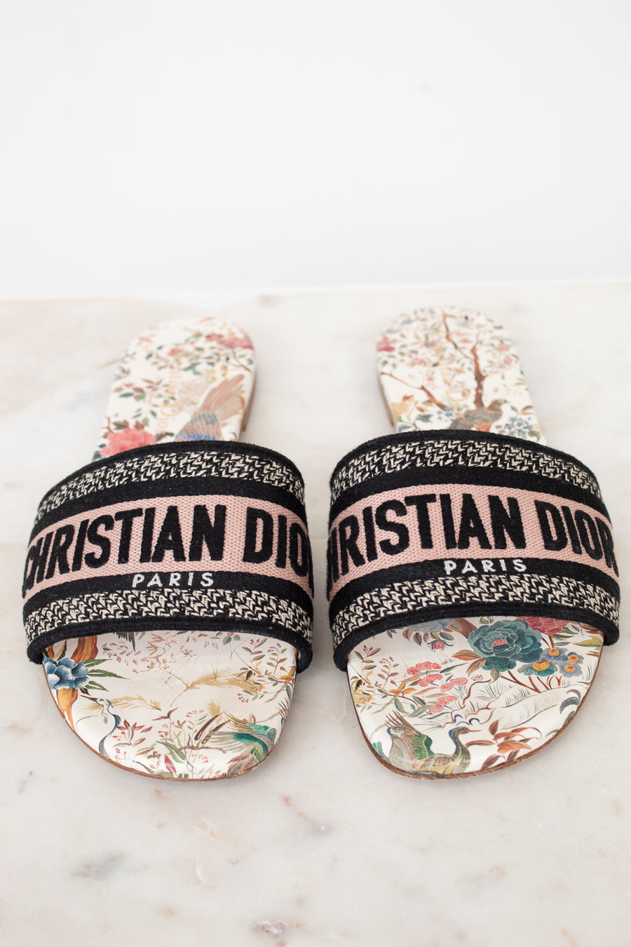 Pair of floral sandals with 'Christian Dior' woven into black/white/pink strap on a marble surface and white background