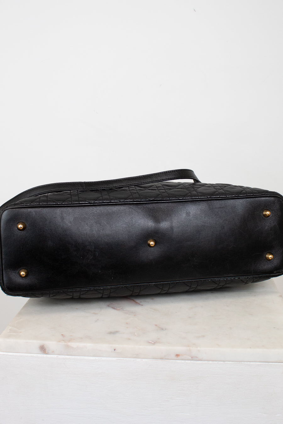 A black leather handbag shown from the bottom, featuring a quilted pattern, gold-tone feet, and a handle, resting on a white marble surface against a white background.