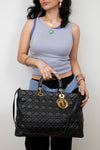A woman holding a black quilted Christian Dior Lady Dior handbag with gold-tone hardware and a signature CD charm, wearing a light blue ribbed tank top and black pants against a plain white background.