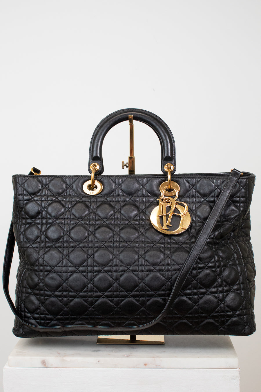 A black quilted leather top-handle handbag with gold-tone hardware, displayed on a white pedestal against a light gray background.