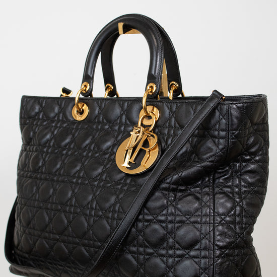 A black quilted leather handbag with gold-tone hardware, featuring dual top handles and a shoulder strap, displayed on a light neutral background.
