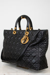 A black quilted leather handbag with gold-tone hardware, featuring dual top handles and a shoulder strap, displayed on a light neutral background.