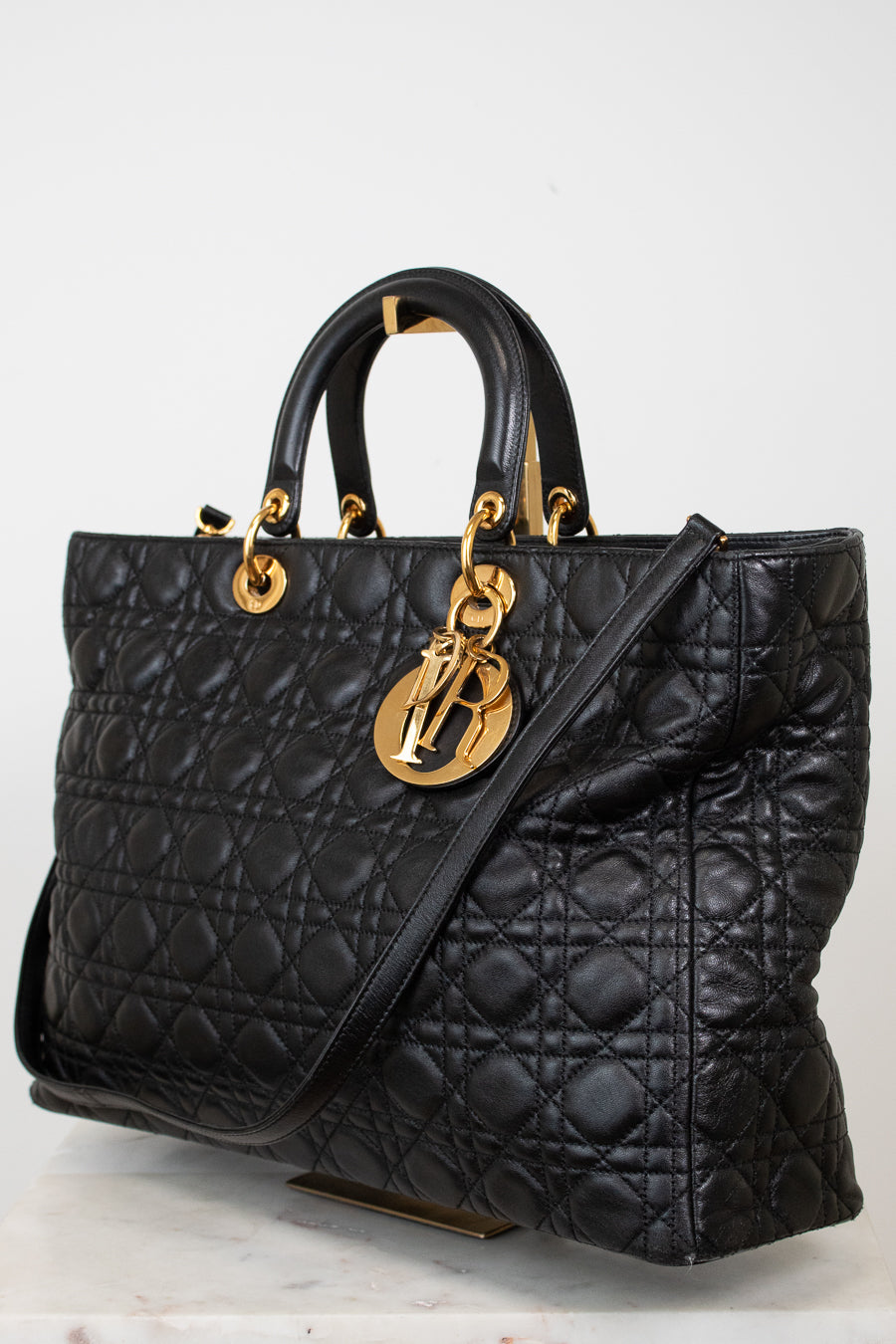 A black quilted leather handbag with gold-tone hardware, featuring dual top handles and a shoulder strap, displayed on a light neutral background.