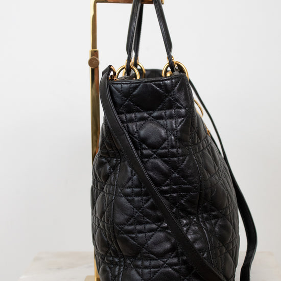 A black quilted leather handbag with gold-tone hardware, displayed on a gold stand against a white background.
