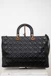 A black quilted leather top-handle satchel bag with gold-tone hardware, displayed on a white marble block against a white background.