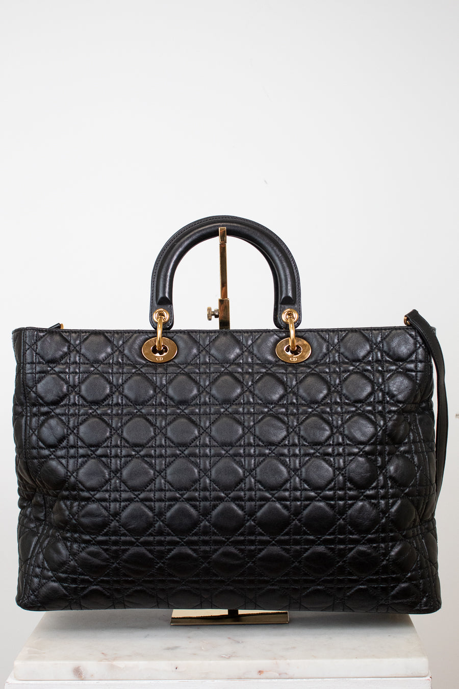A black quilted leather top-handle satchel bag with gold-tone hardware, displayed on a white marble block against a white background.