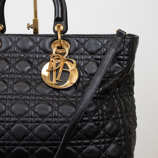 A black quilted leather top-handle handbag with gold-tone hardware, featuring a prominent interlocking 'D' charm and a thin shoulder strap, photographed against a light neutral background.