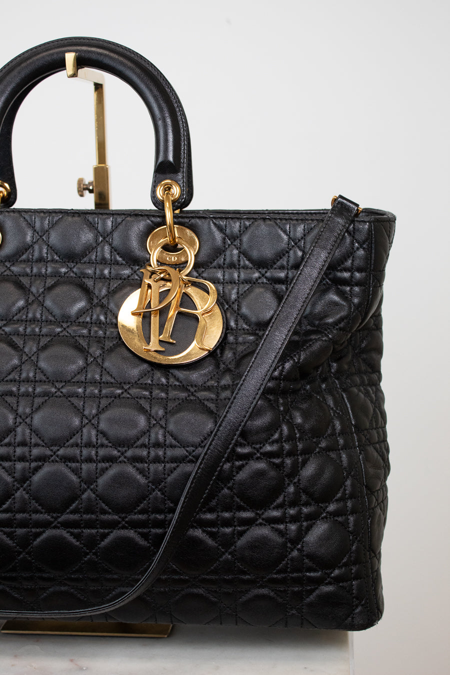 A black quilted leather top-handle handbag with gold-tone hardware, featuring a prominent interlocking 'D' charm and a thin shoulder strap, photographed against a light neutral background.