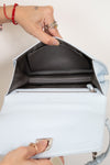 A light blue leather handbag shown open, with a person's hands holding it to display the interior. The bag has a light blue exterior, a dark gray interior lining, and silver-tone hardware.