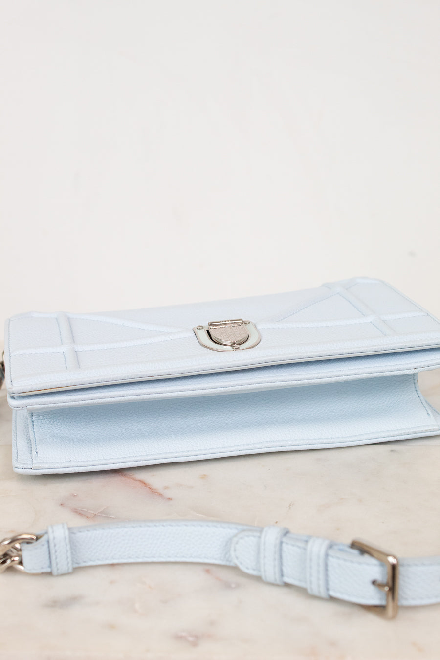 A light blue leather bag with a silver-tone clasp, displayed on a light marble surface with its matching strap in the foreground.