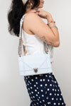 A woman wearing a white sleeveless tank top and navy polka-dot pants carries a light blue structured shoulder bag with a silver chain strap against a light gray background.