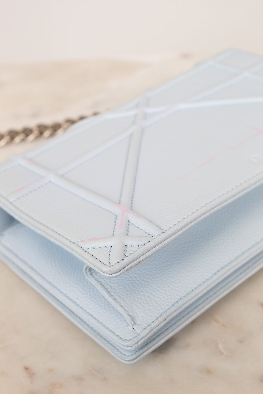 A light blue pebbled leather bag with geometric quilted stitched details, shown on a light marble surface with a silver-tone chain partially visible.