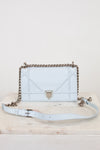 A light blue leather shoulder bag with a chain strap with matching light blue leather strap details, displayed on a marble surface against a white background.