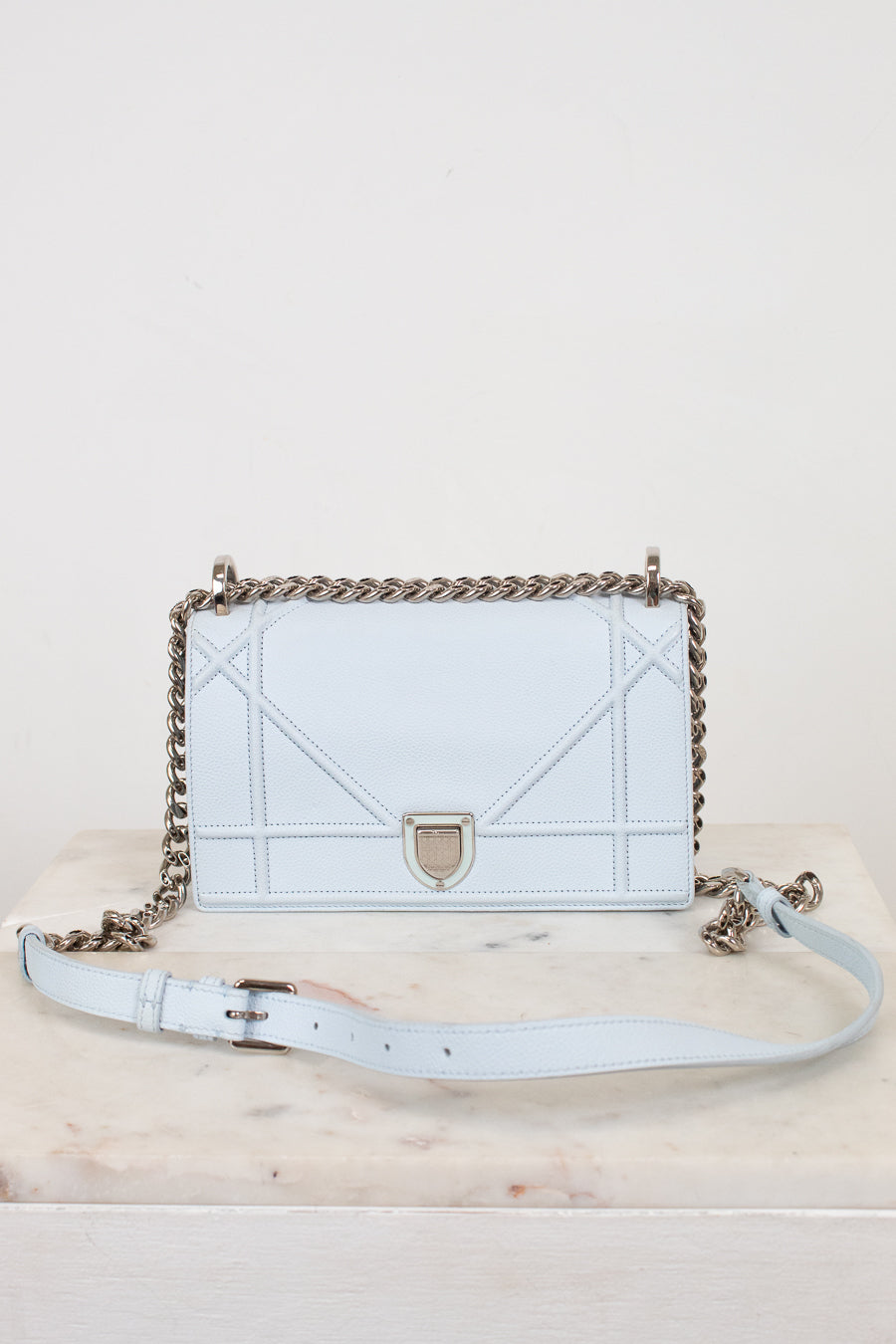 A light blue leather shoulder bag with a chain strap with matching light blue leather strap details, displayed on a marble surface against a white background.