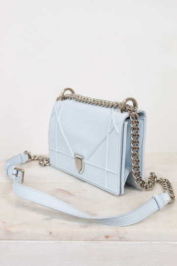 A light blue quilted leather shoulder bag with a silver-tone chain strap and a matching light blue shoulder strap, displayed on a light marble surface against a white background.