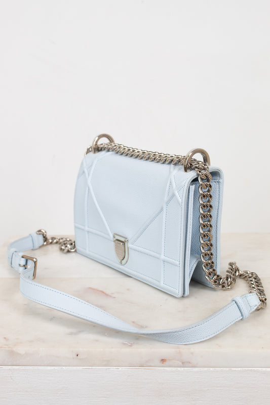 A light blue quilted leather shoulder bag with a silver-tone chain strap and a matching light blue shoulder strap, displayed on a light marble surface against a white background.