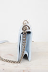 A light blue leather shoulder bag shown from the side, featuring a silver-tone chain strap and a silver-tone metal ring detail, set against a plain white background.
