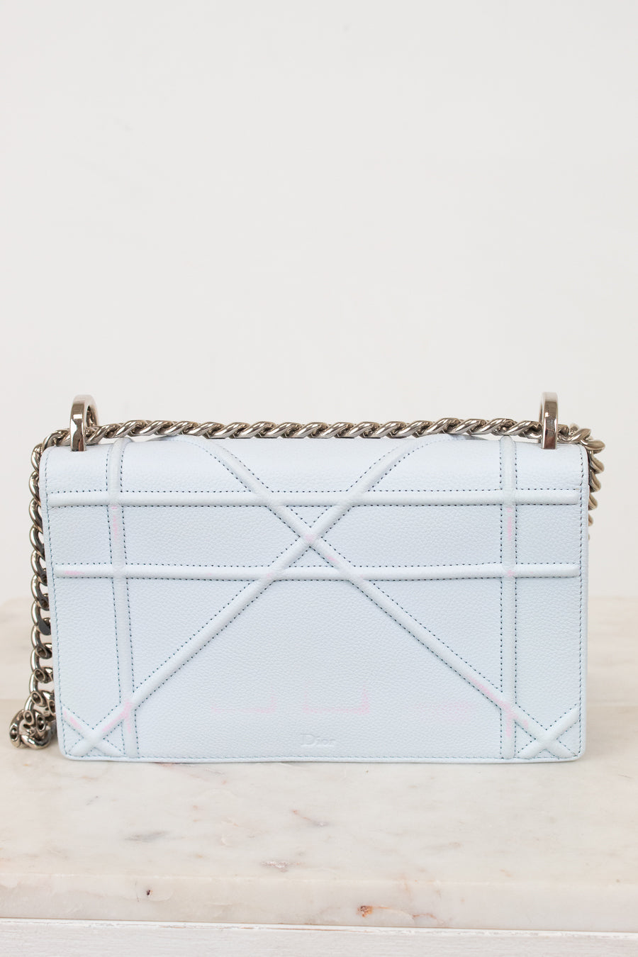 A light blue quilted leather shoulder bag with a geometric quilted pattern and a silver-tone chain strap, resting on a light marble surface against a plain off-white background.