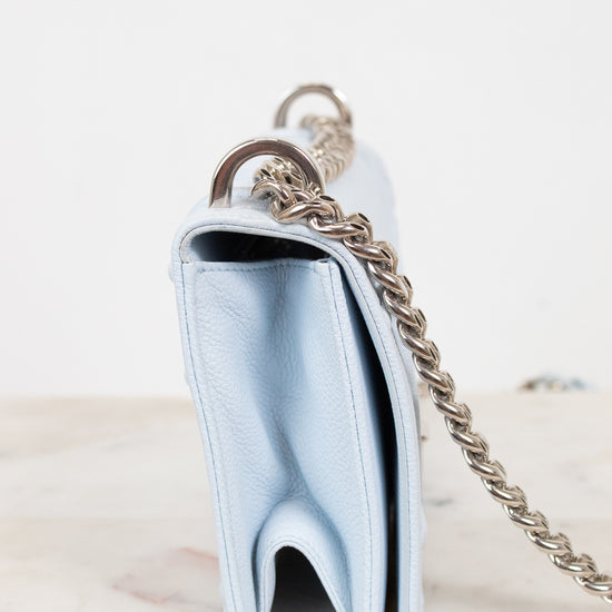 A light blue leather shoulder bag shown from a side profile view, featuring a silver-tone chain strap and a silver-tone metal ring, resting on a light marble surface against a white background.