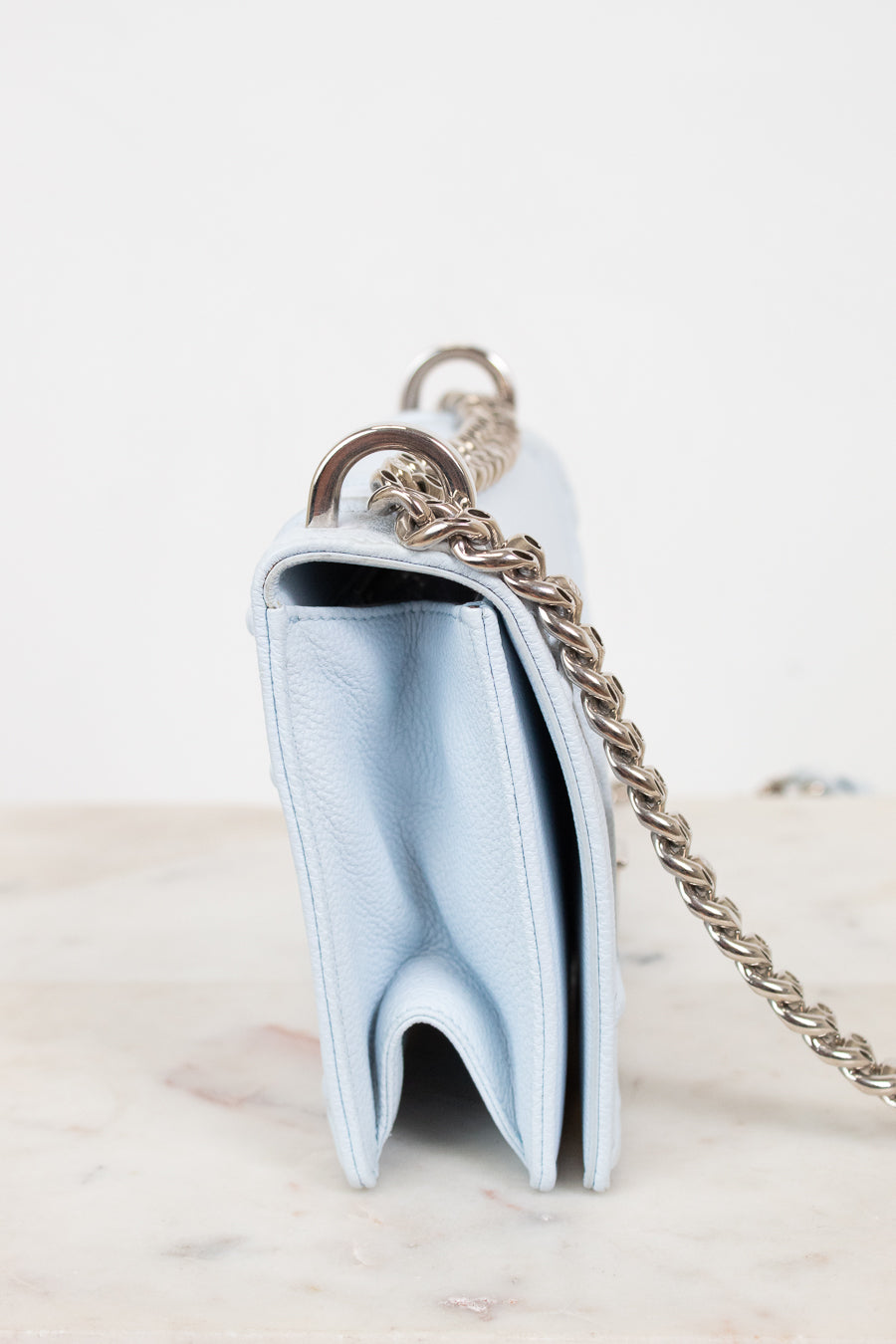 A light blue leather shoulder bag shown from a side profile view, featuring a silver-tone chain strap and a silver-tone metal ring, resting on a light marble surface against a white background.