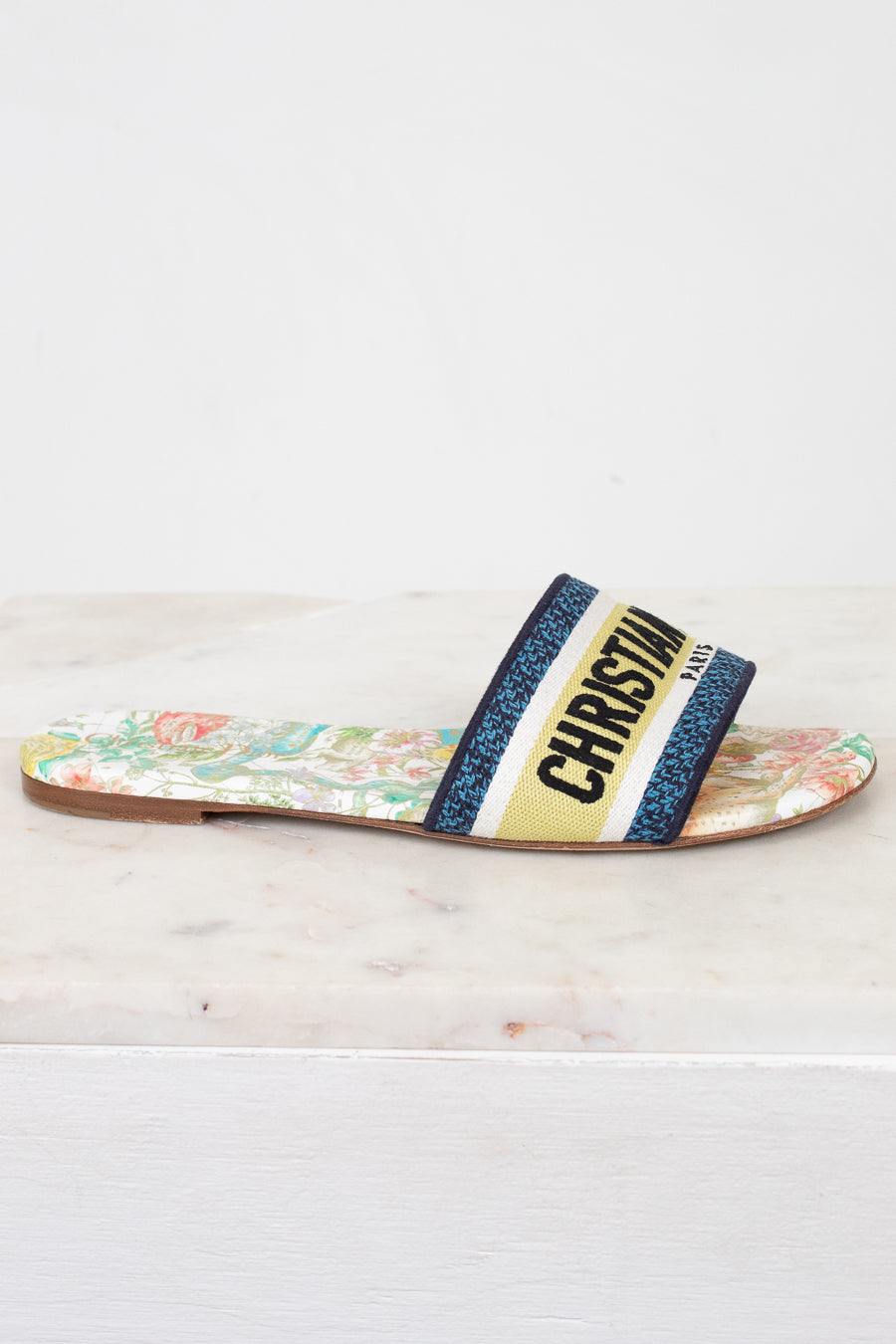 Side view of floral sandals with 'Christian Dior' blue/black/white/chartreuse strap on a marble surface