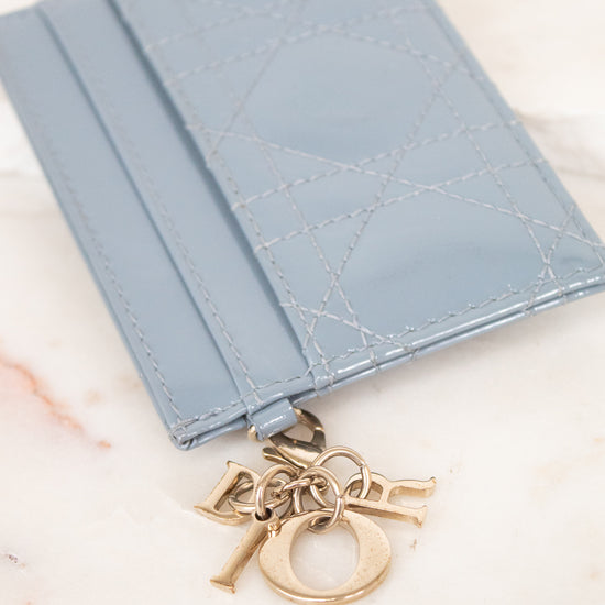 A light blue leather card holder with a signature Dior Oblique quilted pattern and a gold-tone Dior charm, displayed on a light marble surface.