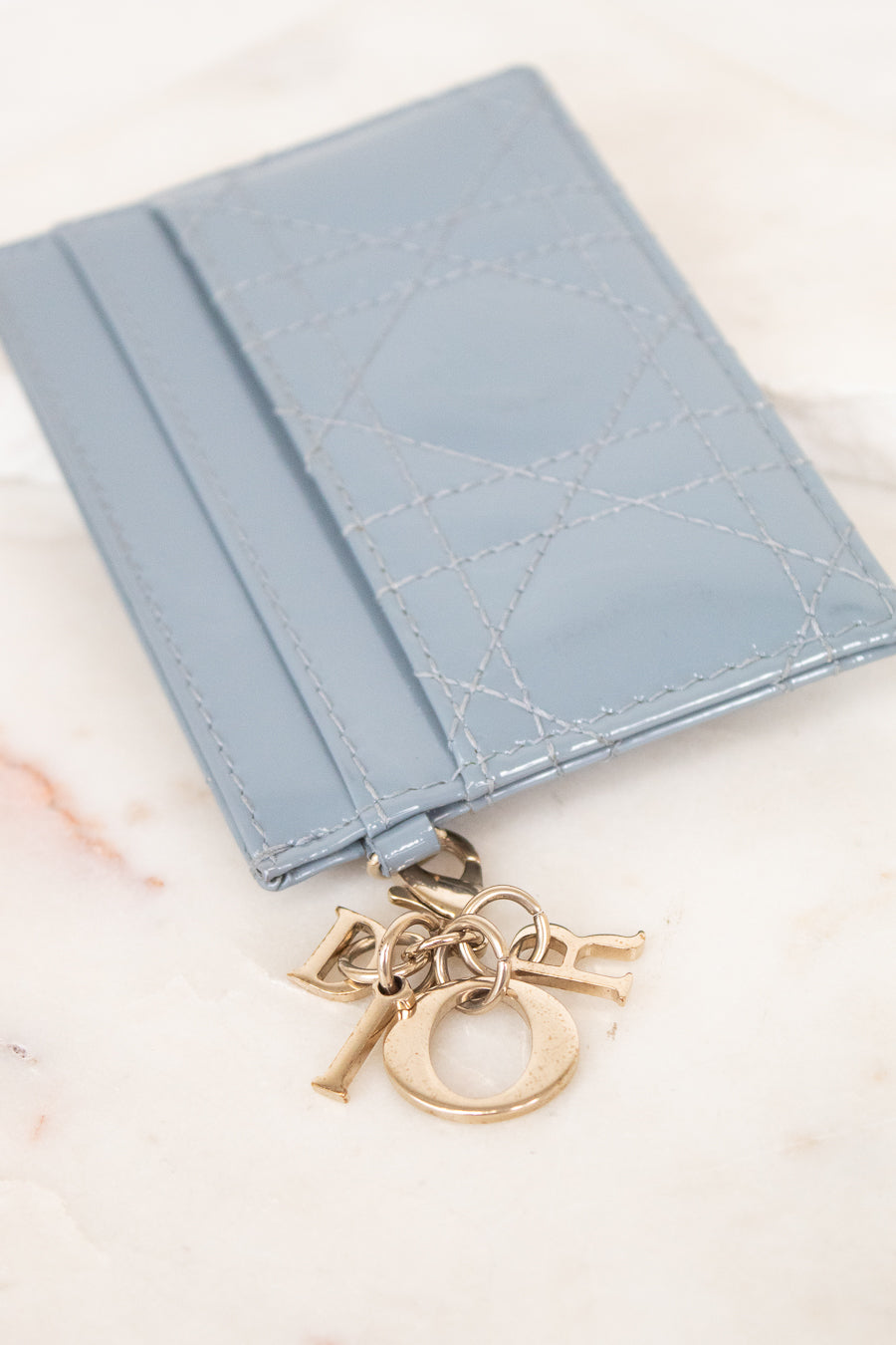 A light blue leather card holder with a signature Dior Oblique quilted pattern and a gold-tone Dior charm, displayed on a light marble surface.