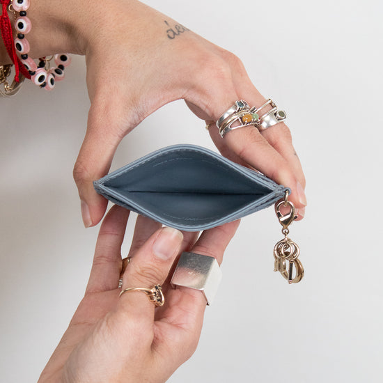 A close-up of a person's hands holding an open slim card holder wallet with a small gold charm attached, set against a plain light gray background.