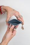 A close-up of a person's hands holding an open slim card holder wallet with a small gold charm attached, set against a plain light gray background.