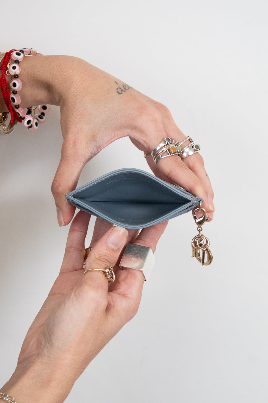 A close-up of a person's hands holding an open slim card holder wallet with a small gold charm attached, set against a plain light gray background.