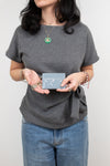 A woman wearing a heather gray short-sleeve t-shirt with a side tie detail, paired with blue jeans. She is holding a small light blue card holder in front of her torso.