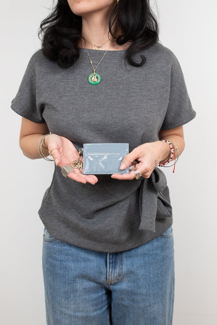 A woman wearing a heather gray short-sleeve t-shirt with a side tie detail, paired with blue jeans. She is holding a small light blue card holder in front of her torso.