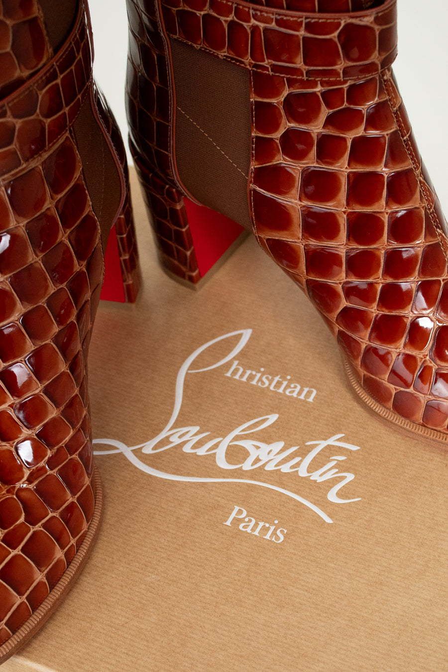 Brown crocodile-patterned high-heeled shoes on a beige surface with Christian Louboutin branding.