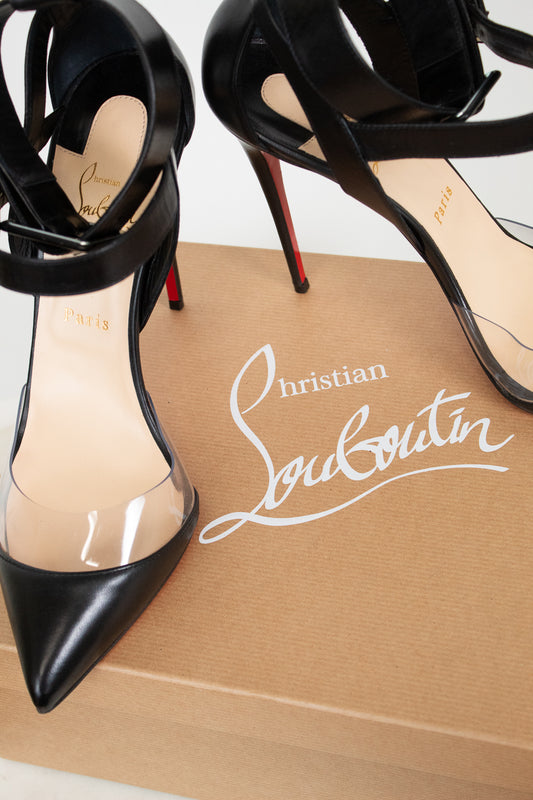 A pair of Christian Louboutin black leather high-heeled sandals with transparent PVC side panels and red soles, displayed on top of a brown shoebox with the white Christian Louboutin logo.