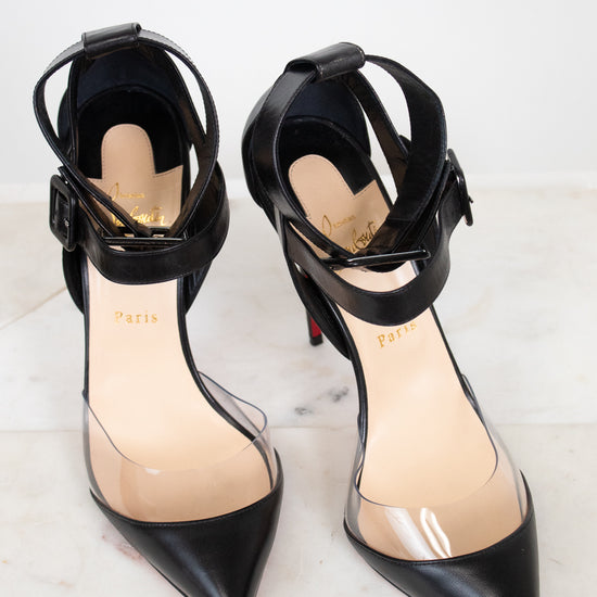 A pair of black pointed-toe high heels with PVC panel and ankle straps, shown on a light marble surface against a white background.