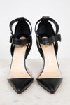 A pair of black pointed-toe high heels with clear side panels and ankle straps, shown on a light marble surface against a white background.