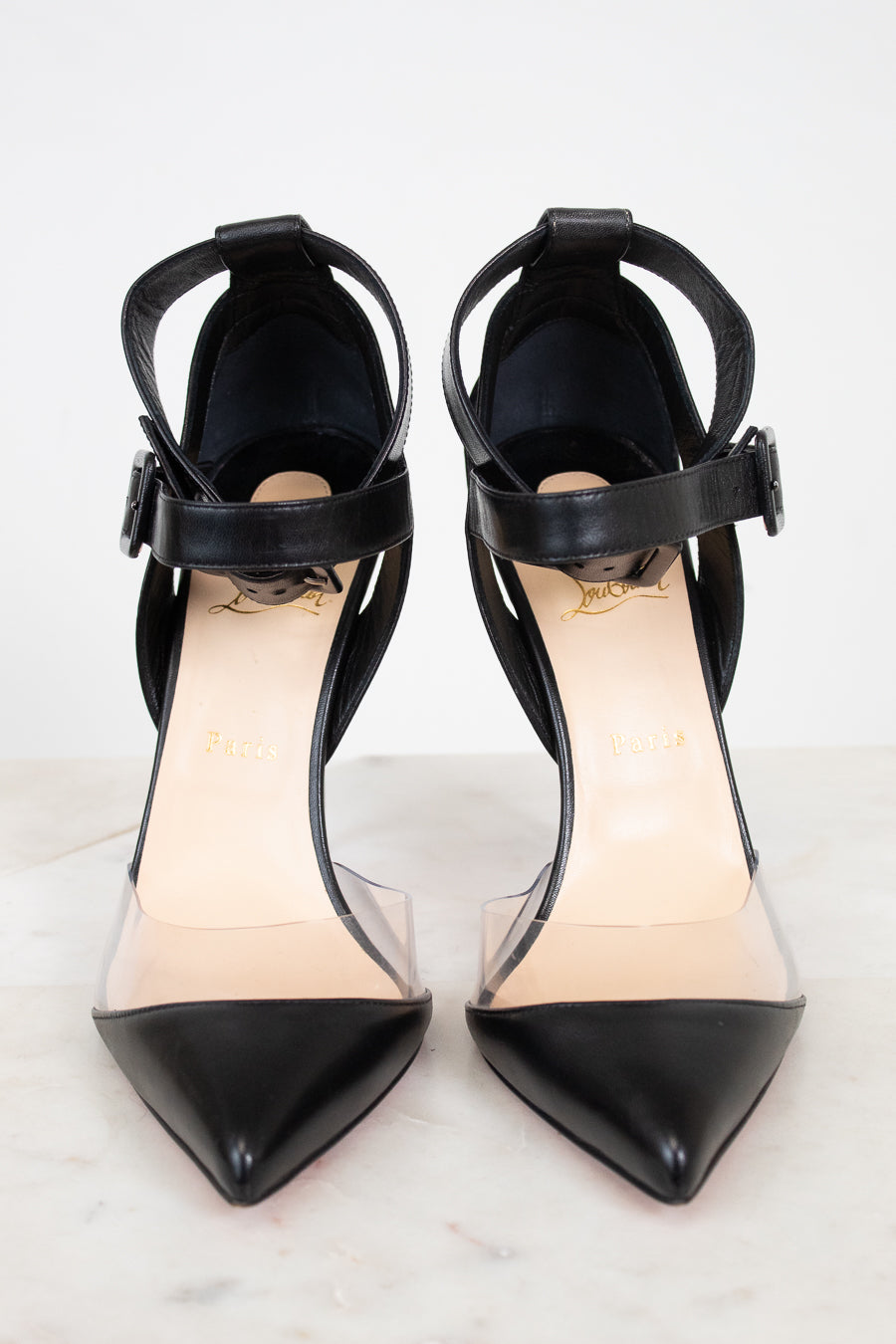 A pair of black pointed-toe high heels with clear side panels and ankle straps, shown on a light marble surface against a white background.