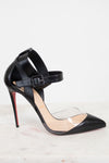A black leather high-heeled pump with a pointed toe, a clear PVC toe strip, and a buckle strap around the ankle, set against a white background with a signature red sole.