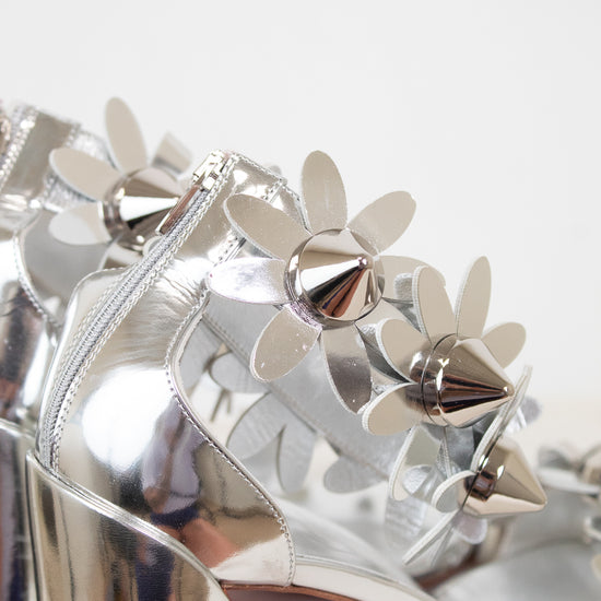 Silver high-heeled shoes with floral studded embellishments on a light background