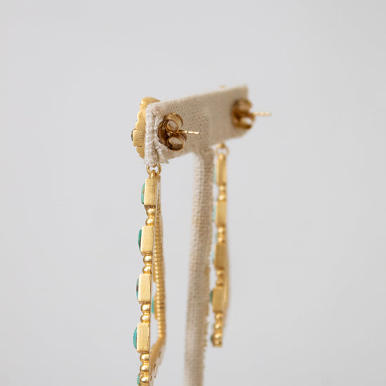 A pair of gold dangle earrings with small turquoise blue stones, displayed on a cream fabric jewelry stand against a light gray background.