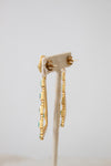 A pair of gold dangle earrings with small turquoise blue stones, displayed on a cream fabric jewelry stand against a light gray background.