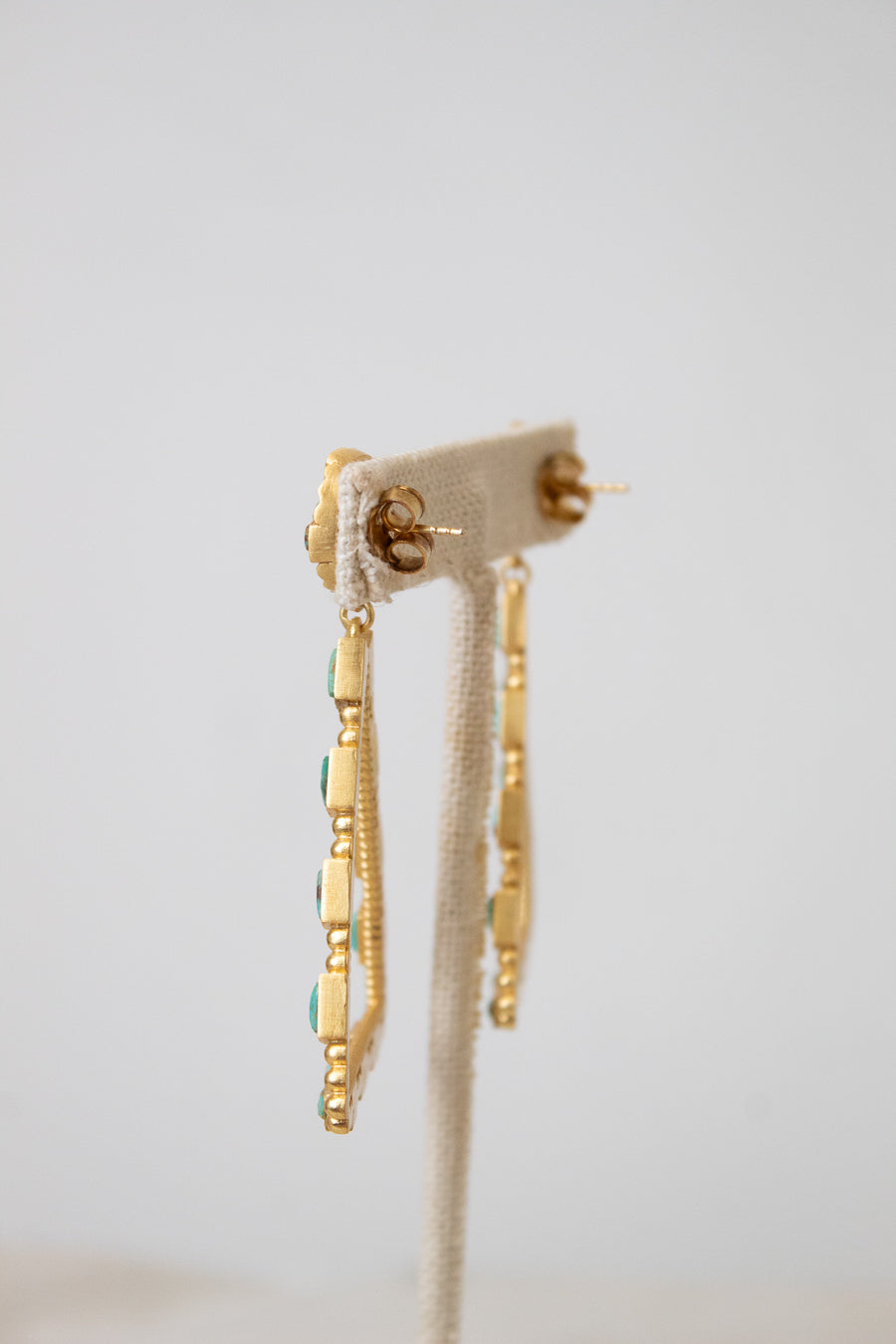 A pair of gold dangle earrings with small turquoise blue stones, displayed on a cream fabric jewelry stand against a light gray background.