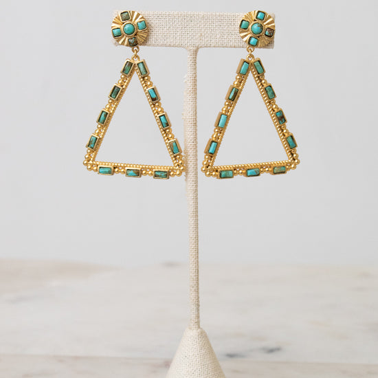 Gold triangle dangle earrings with turquoise blue rectangular stones and round turquoise coin posts, displayed on a cream textured stand.