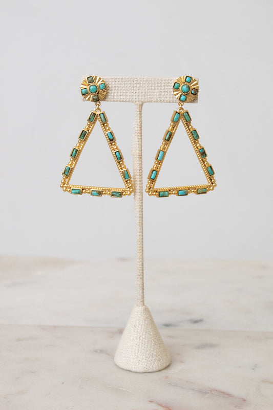 Gold triangle dangle earrings with turquoise blue rectangular stones and round turquoise coin posts, displayed on a cream textured stand.