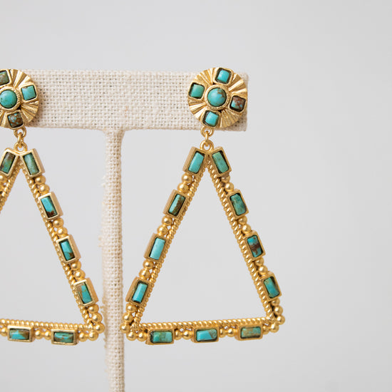 Gold triangular drop earrings with turquoise-colored rectangular stones, displayed on a beige fabric jewelry stand against a light gray background.