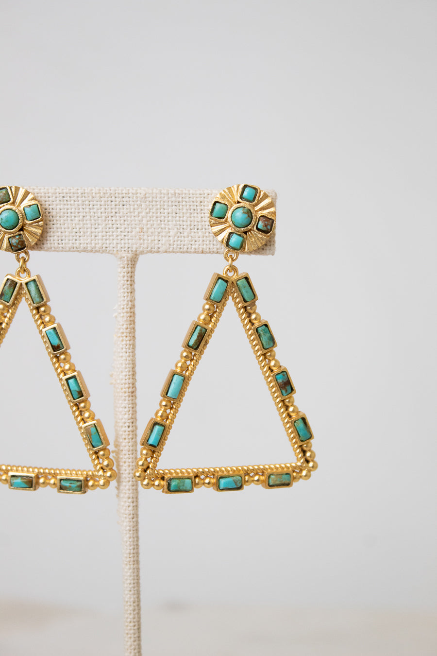 Gold triangular drop earrings with turquoise-colored rectangular stones, displayed on a beige fabric jewelry stand against a light gray background.