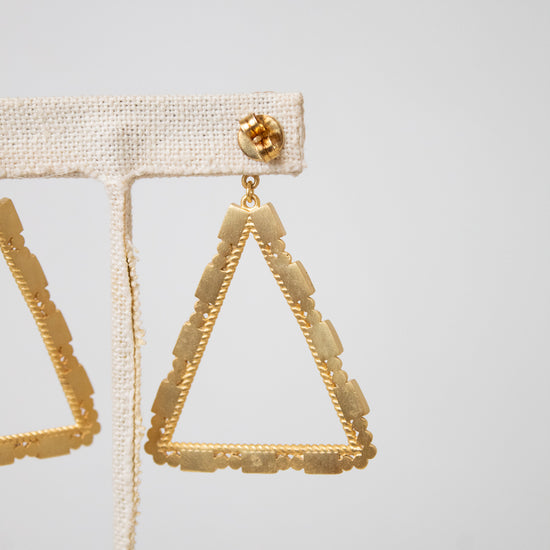 Gold triangular dangle earrings with a textured beaded border, displayed on a beige fabric jewelry stand against a light gray background.